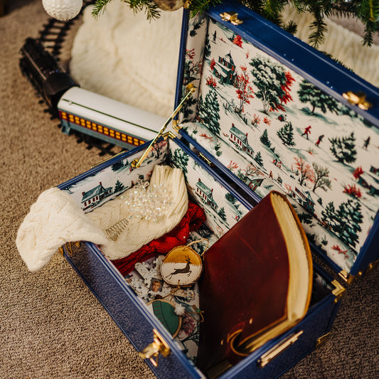 Open trunk with Christmas-themed interior, including a book and decorative items, with a Christmas tree and train in the background.
