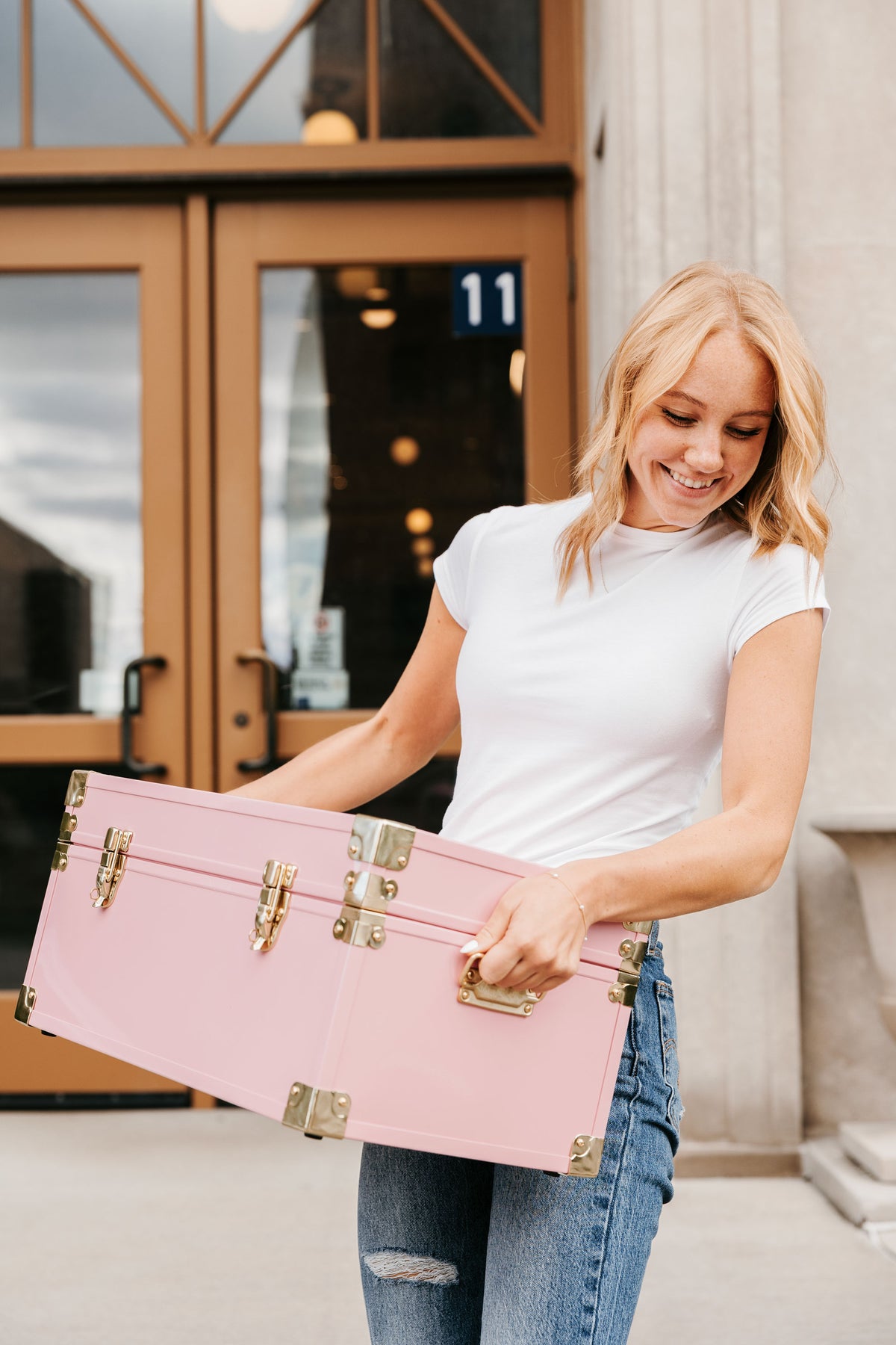 A woman in a white t-shirt and blue jeans holds a light pink vintage-style hard case Momento trunk with gold-tone metal hardware, standing in front of a building entrance.