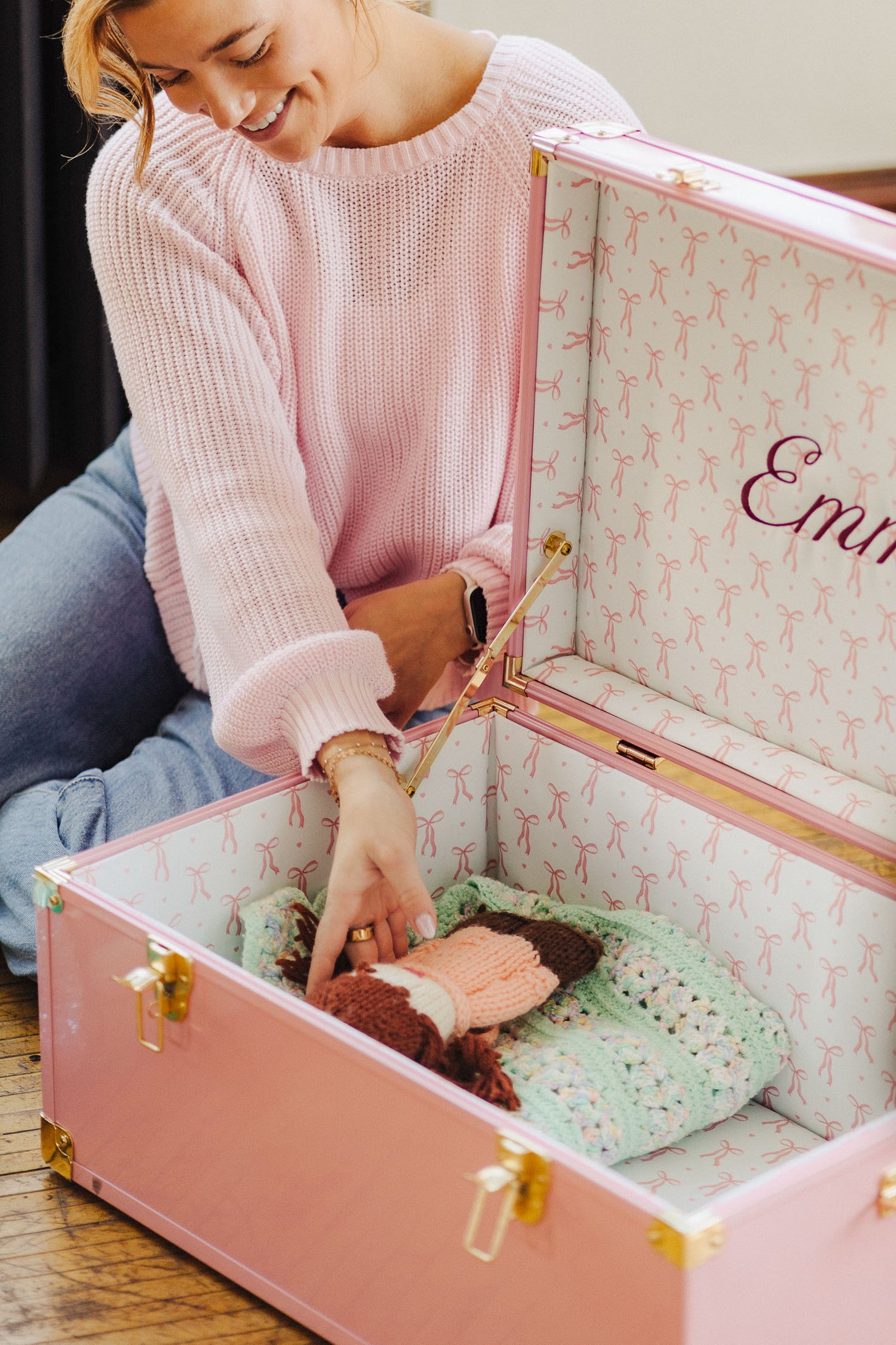 Woman setting a baby toy into a pink keepsake trunk, sitting on a wooden floor.