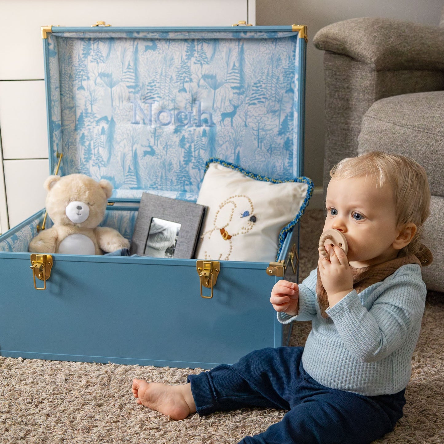 Child sitting next to a blue keepsake trunk filled with sentimental toys and baby items.