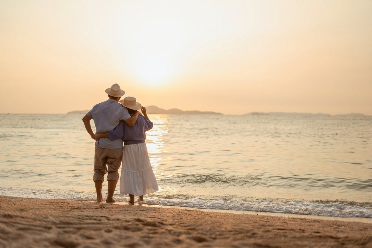 A couple standing on a sandy beach at sunset, facing the ocean. The man wears a light blue shirt, beige pants, and a white hat, while the woman wears a purple top, a long white skirt, and a white hat.