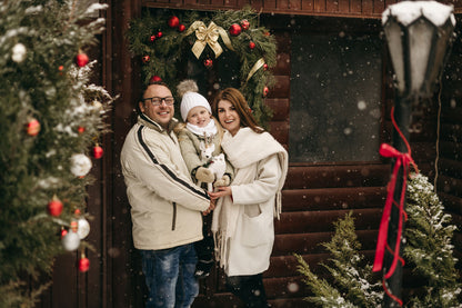 Family of three in winter clothing standing in front of a decorated wooden cabin with Christmas trees.